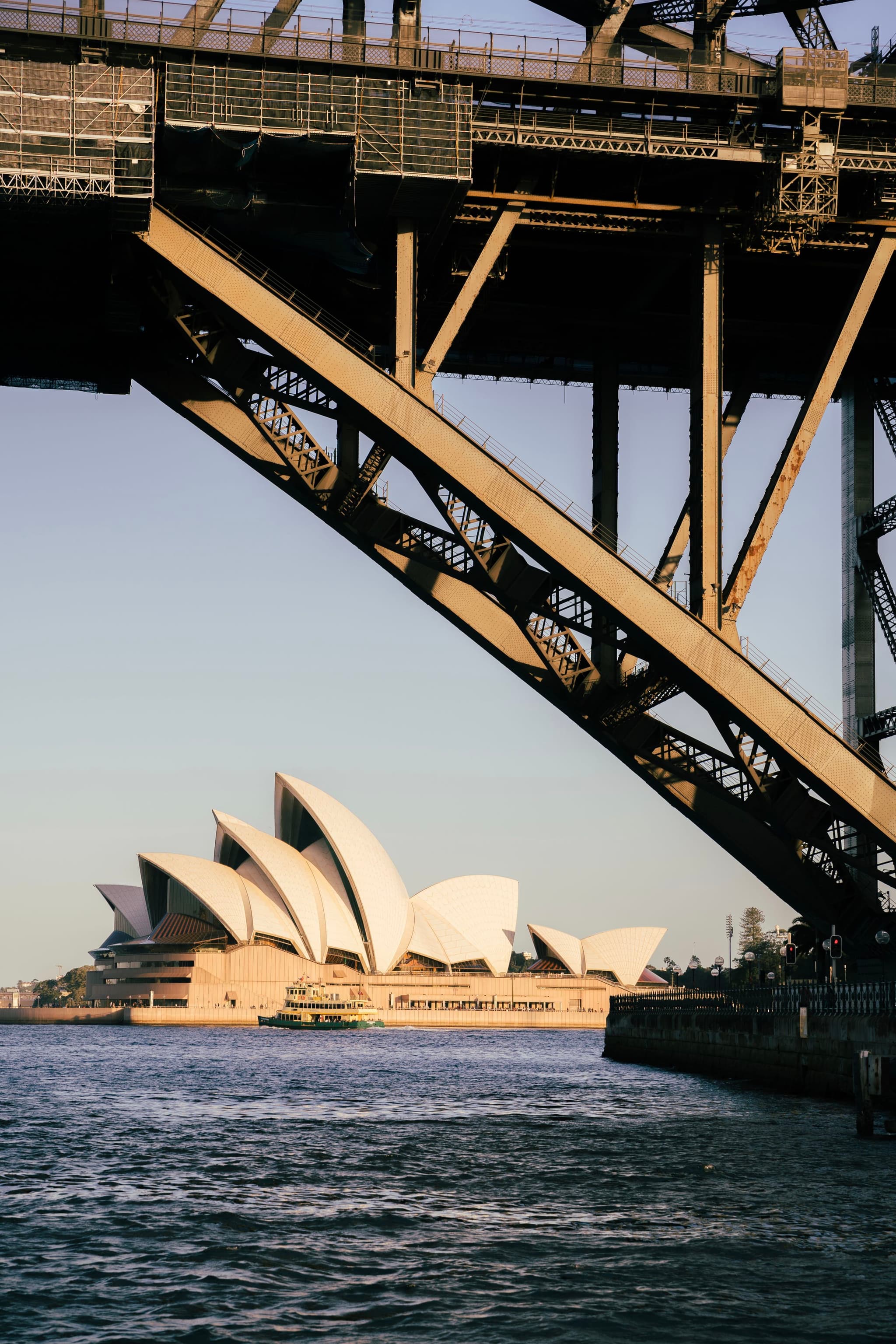 The Sydney Opera House viewed through the structure of the Sydney Harbour Bridge, with water in the foreground
