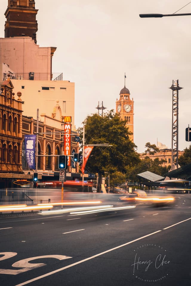 A city street scene with blurred motion from passing vehicles, featuring historic buildings and a clock tower in the background