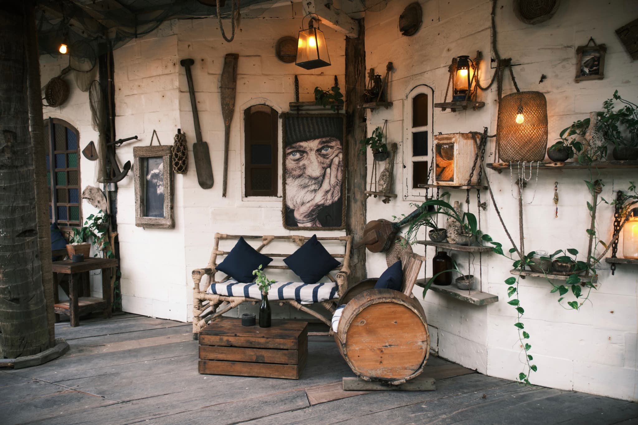 A cozy, rustic interior with a wooden bench, cushions, and various decorative items like oars, lanterns, and plants adorning the walls. A large portrait and a log table add to the eclectic decor