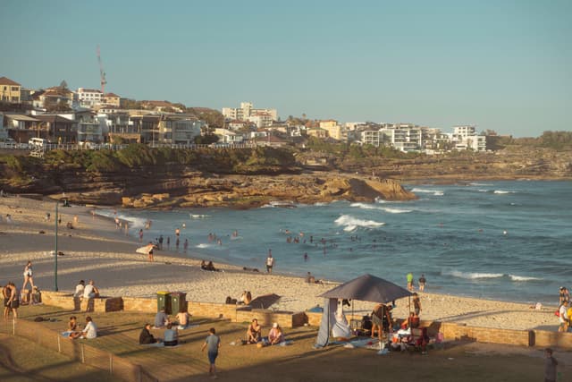 A coastal scene with a sandy beach, people relaxing under a tent, and waves crashing on the shore. Buildings are visible on the cliffs in the background under a clear sky