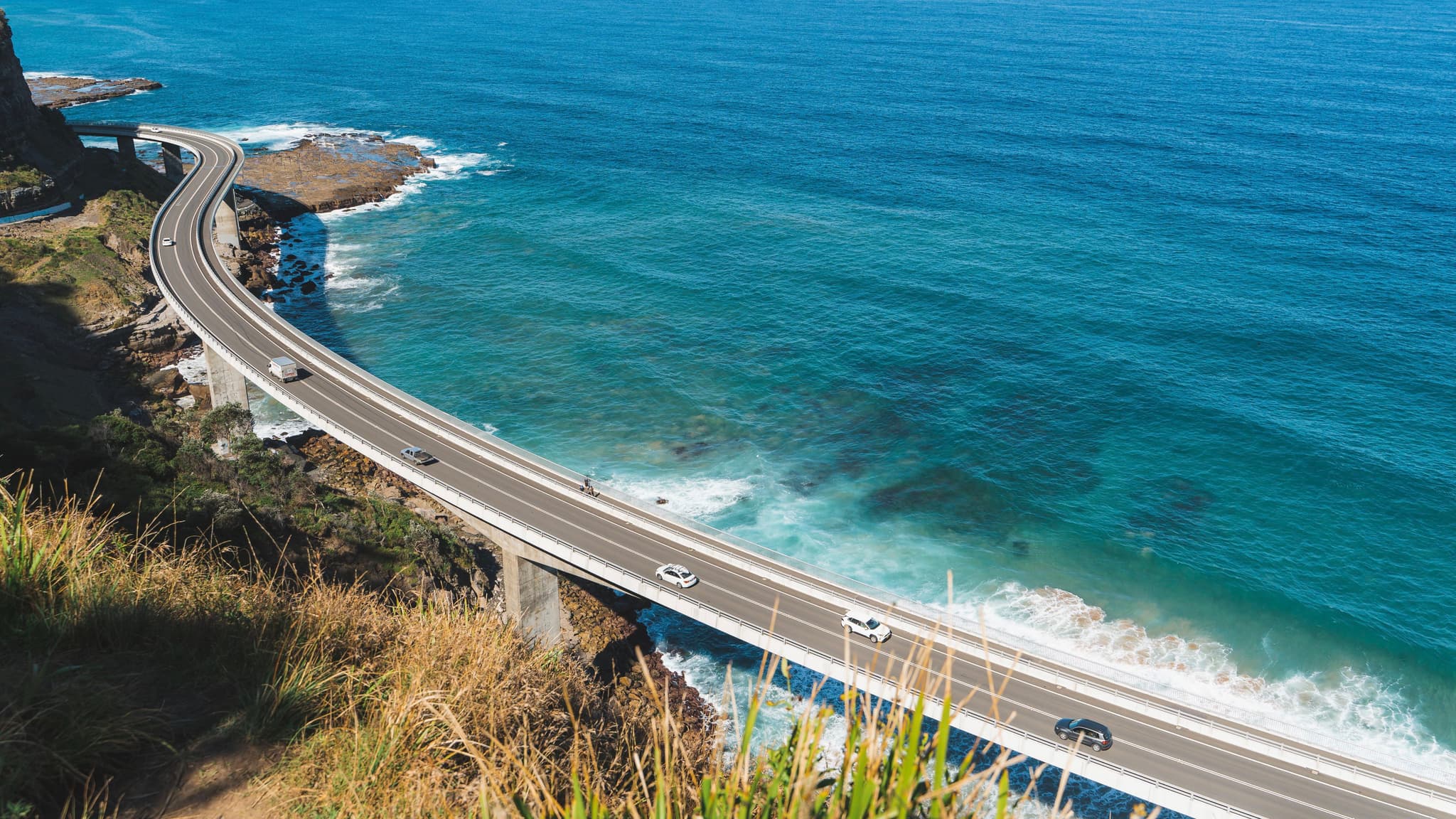 A coastal highway curves along the edge of a cliff, with cars traveling on it. The ocean is a vibrant blue, and waves crash against the shore below. Vegetation is visible in the foreground