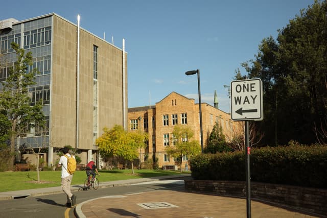 A street scene with a one-way sign, a cyclist, and a pedestrian. Modern and older buildings are in the background, surrounded by trees and greenery under a clear blue sky