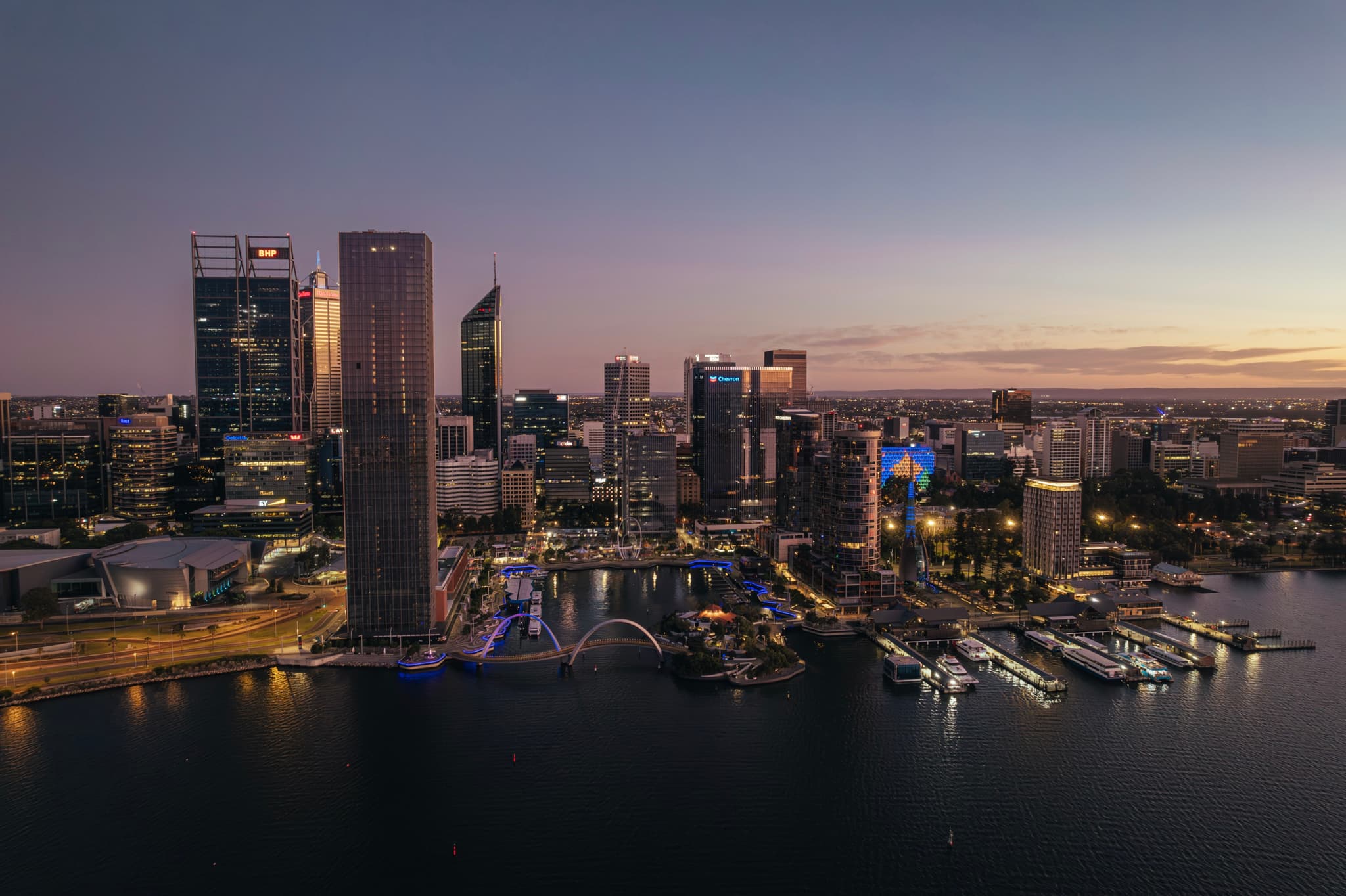 A city skyline at dusk with illuminated skyscrapers and a waterfront, reflecting lights on the water