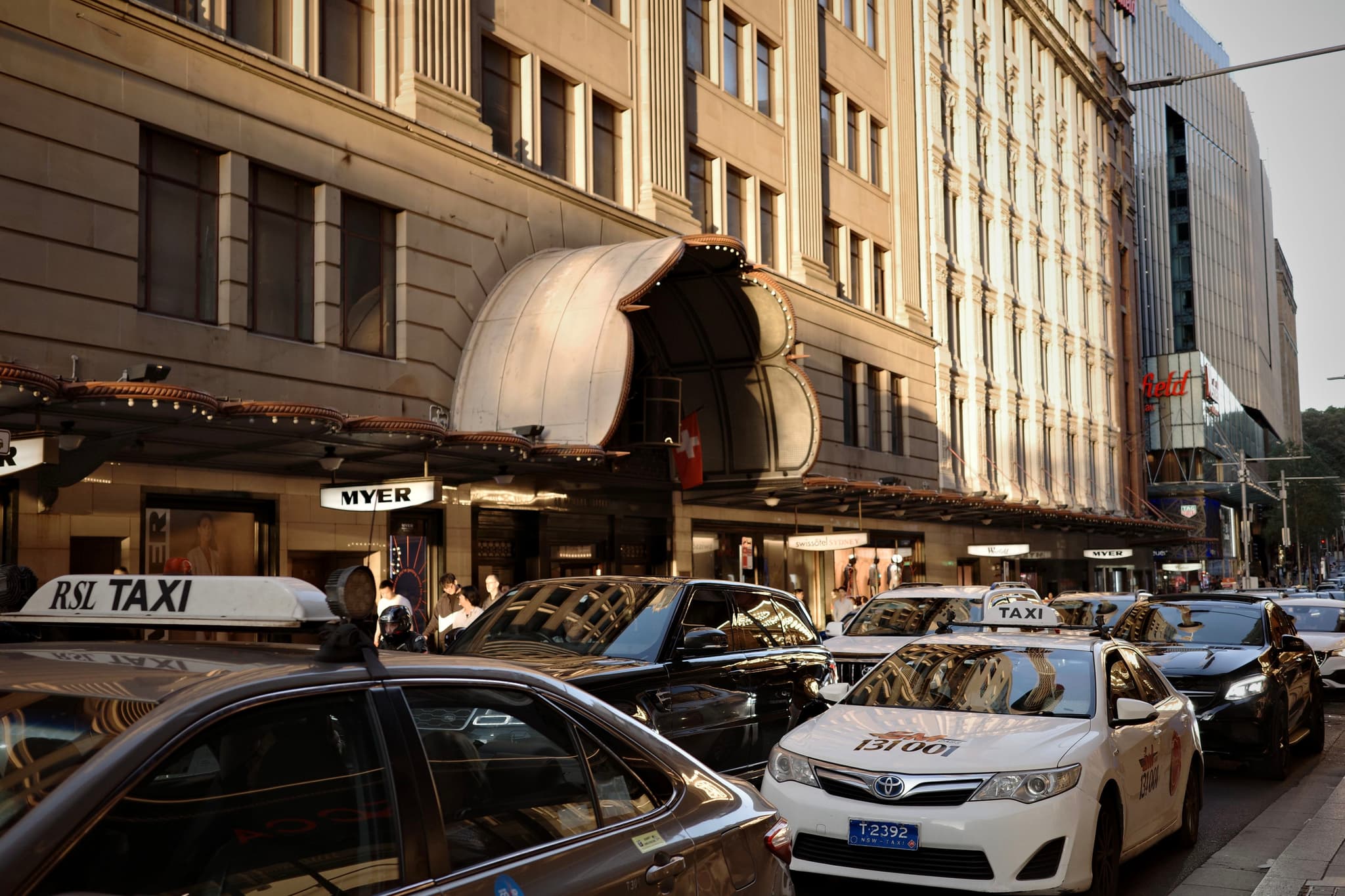 A busy city street with several taxis lined up in front of a large building, featuring a prominent awning and bustling pedestrian activity