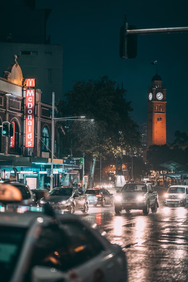 A rainy city street at night with cars, a McDonald's sign, and a clock tower in the background