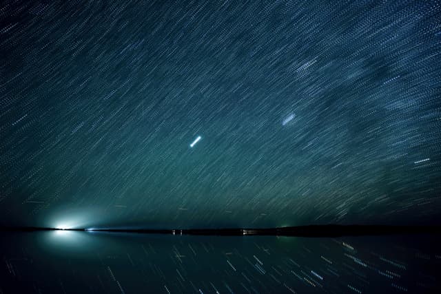 A long-exposure photograph capturing star trails in the night sky above a reflective body of water, with a bright light source on the horizon