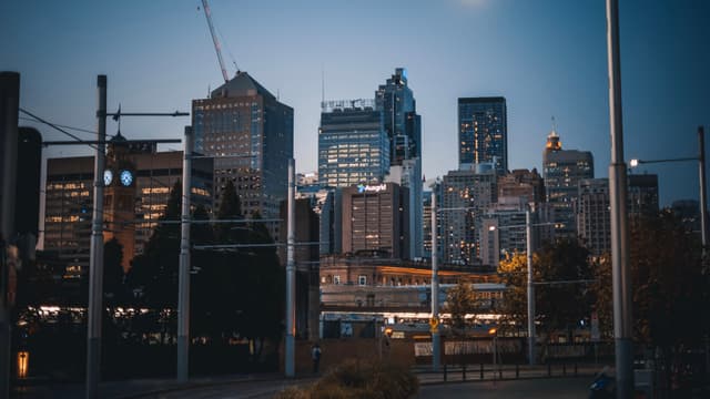 A city skyline at dusk with tall buildings and a clear sky, featuring some trees and streetlights in the foreground