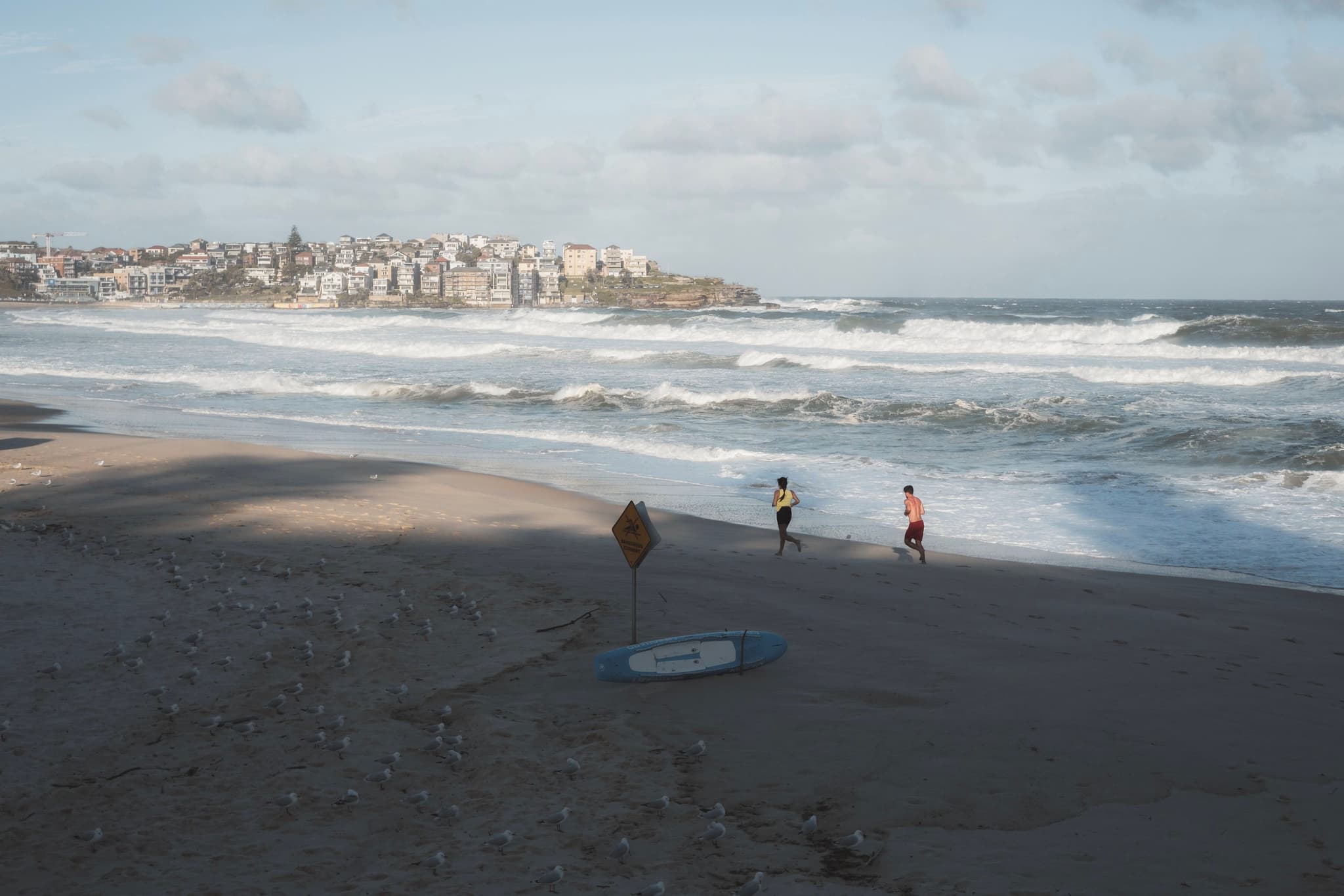 A beach scene with two people jogging along the shoreline, waves crashing onto the sand, and a distant view of buildings on a headland under a cloudy sky