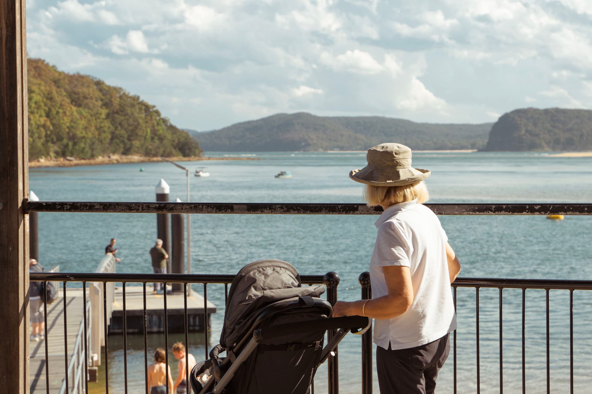 A person with a hat stands by a railing, pushing a stroller, overlooking a scenic body of water with hills in the background
