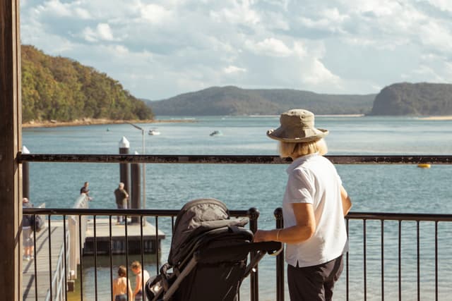 A person with a hat stands by a railing, pushing a stroller, overlooking a scenic body of water with hills in the background