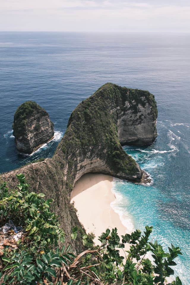 Aerial view of a coastal landscape with a prominent rock formation resembling a dinosaur, surrounded by turquoise waters and a sandy beach, with lush greenery in the foreground
