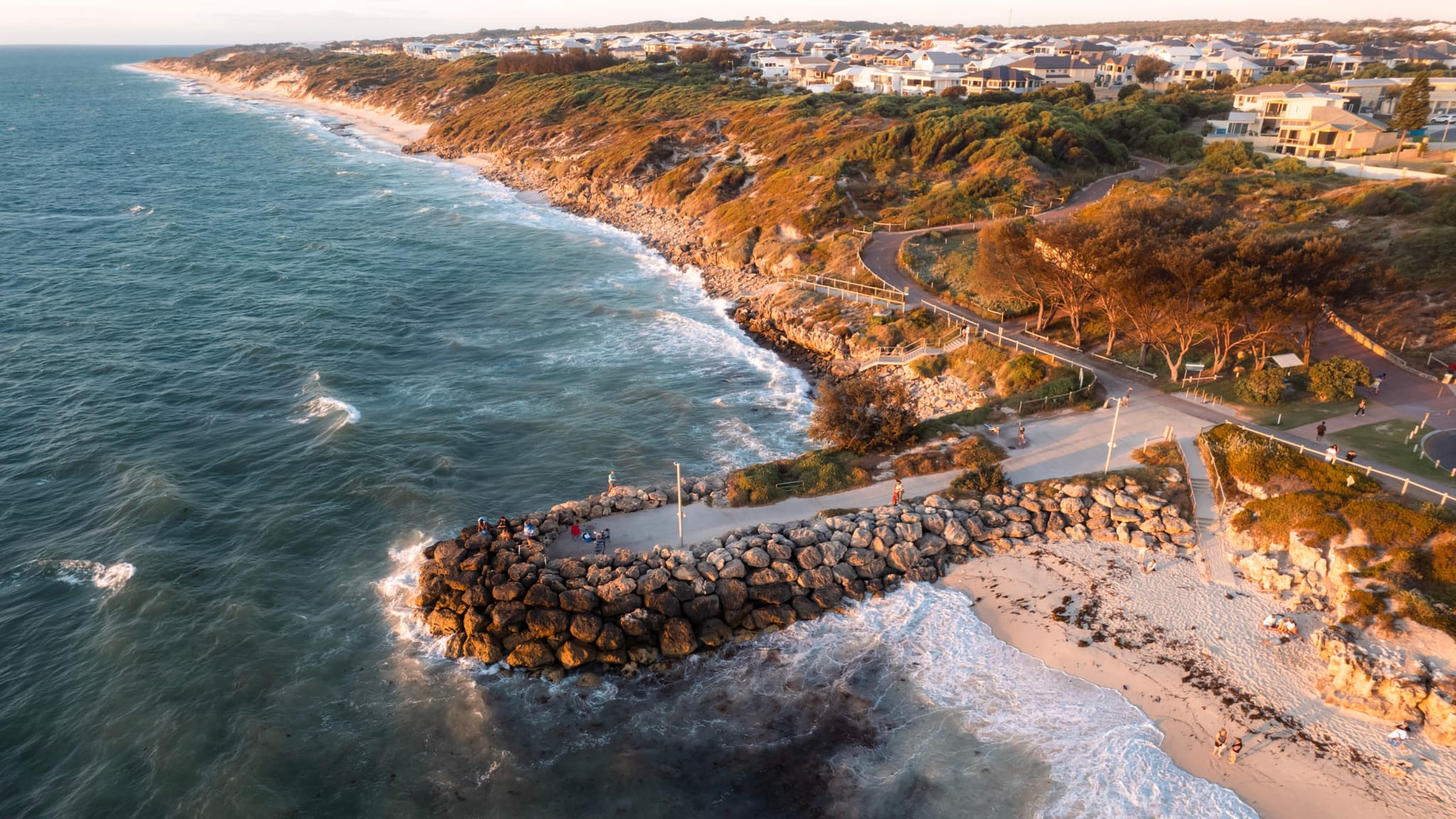 A coastal landscape featuring a rocky shoreline with waves crashing against it, a jetty extending into the sea, and a backdrop of cliffs and buildings under a clear sky