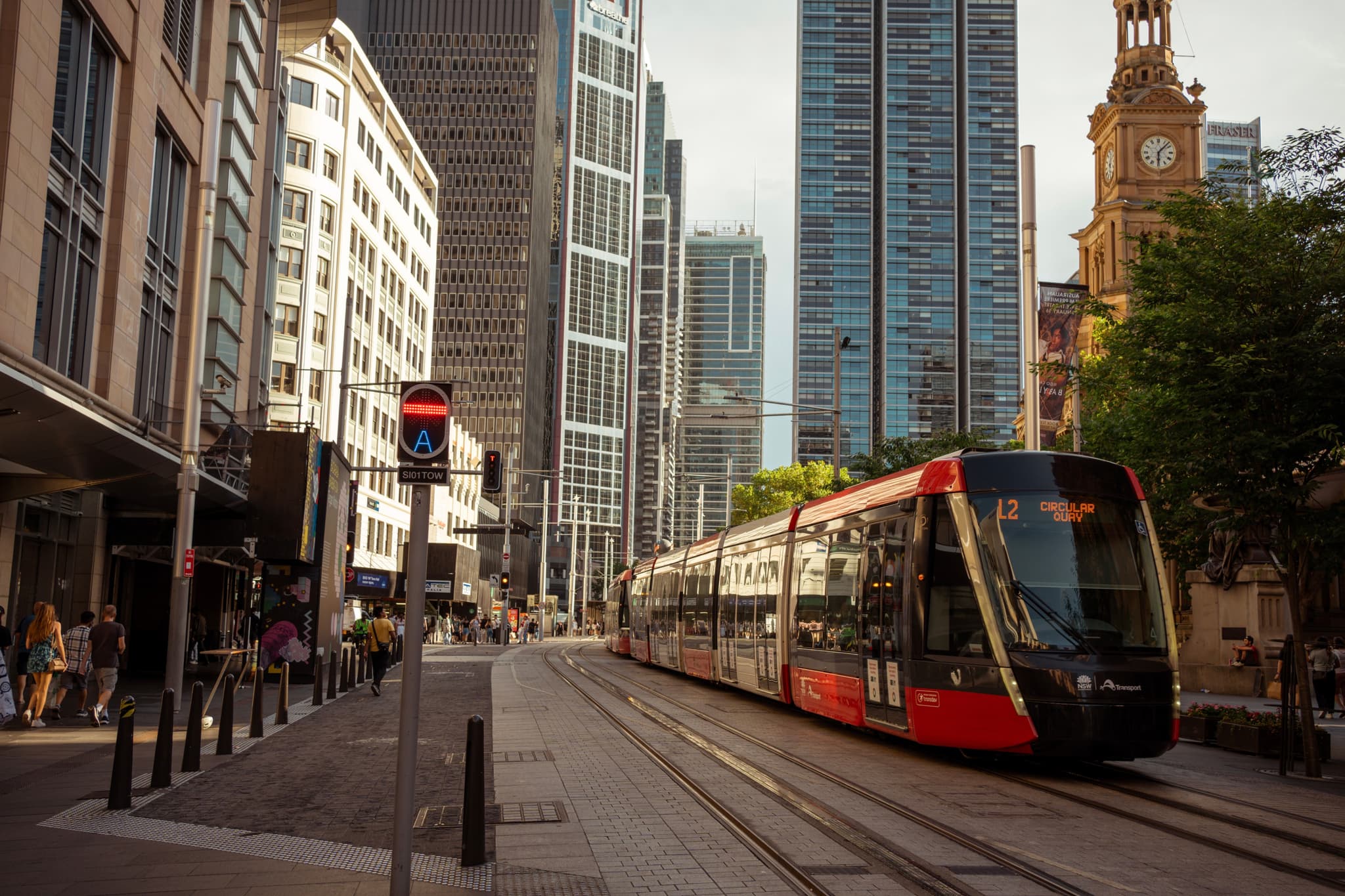 A modern tram travels through a city street flanked by tall buildings, with pedestrians walking along the sidewalks