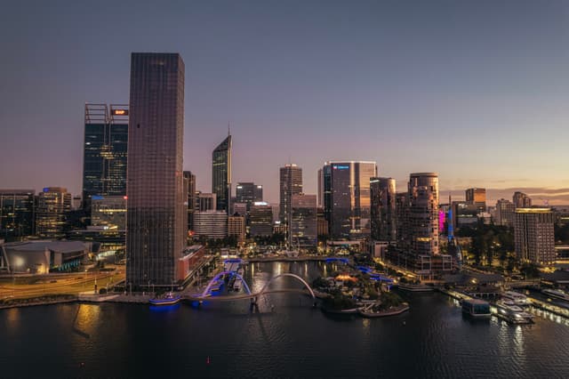 A city skyline at dusk with tall buildings reflecting on a body of water, illuminated by artificial lights