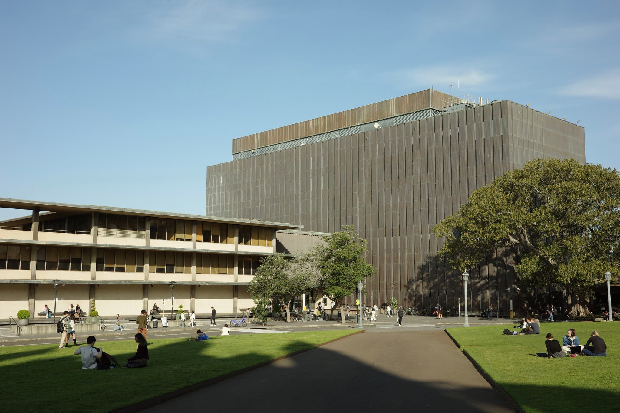 A large, modern building with a grid-like facade stands next to a smaller, elongated structure. People are sitting on the grass and walking along a paved path under a clear blue sky
