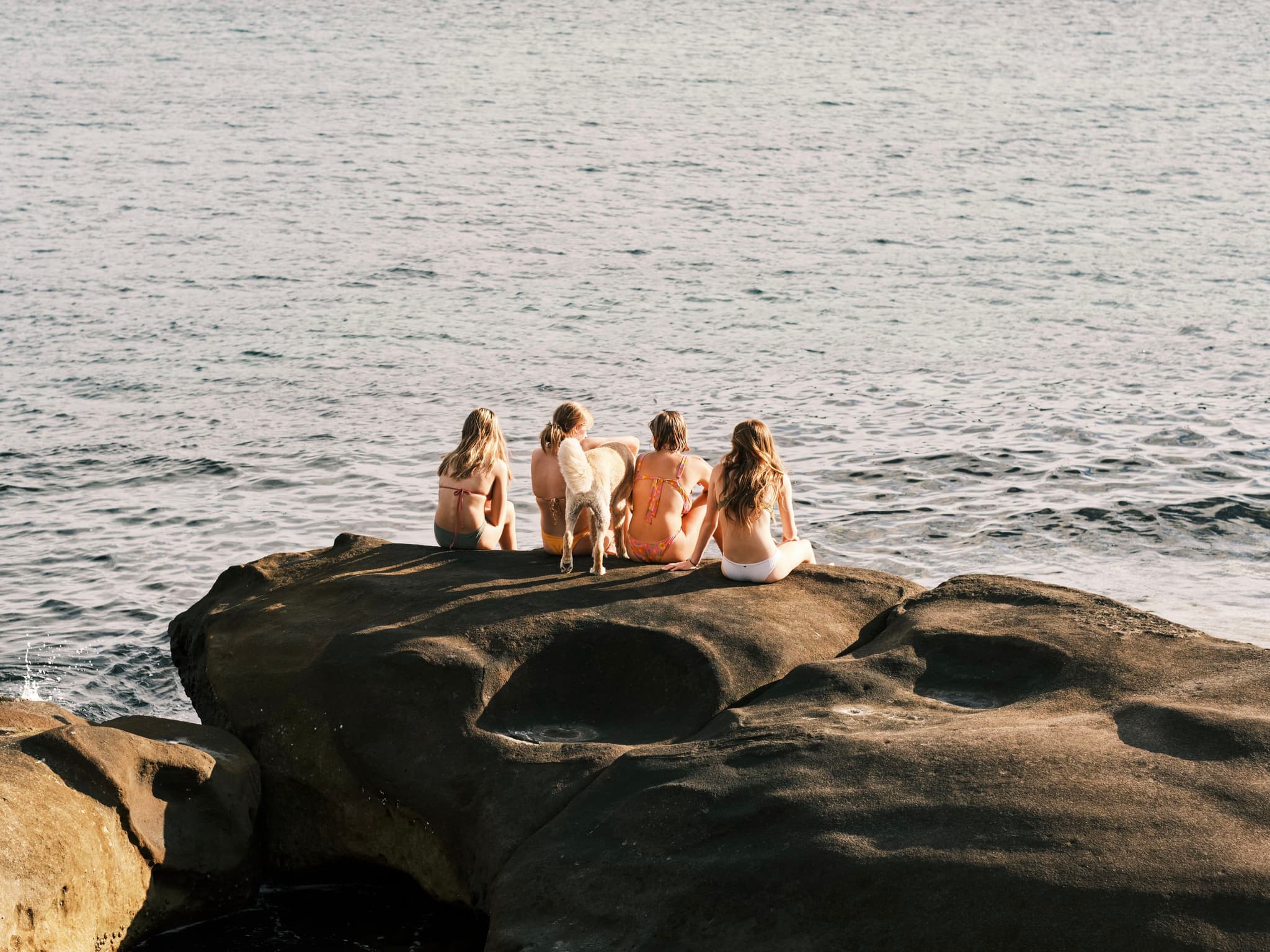 Four people sitting on a large rock by the ocean, facing the water