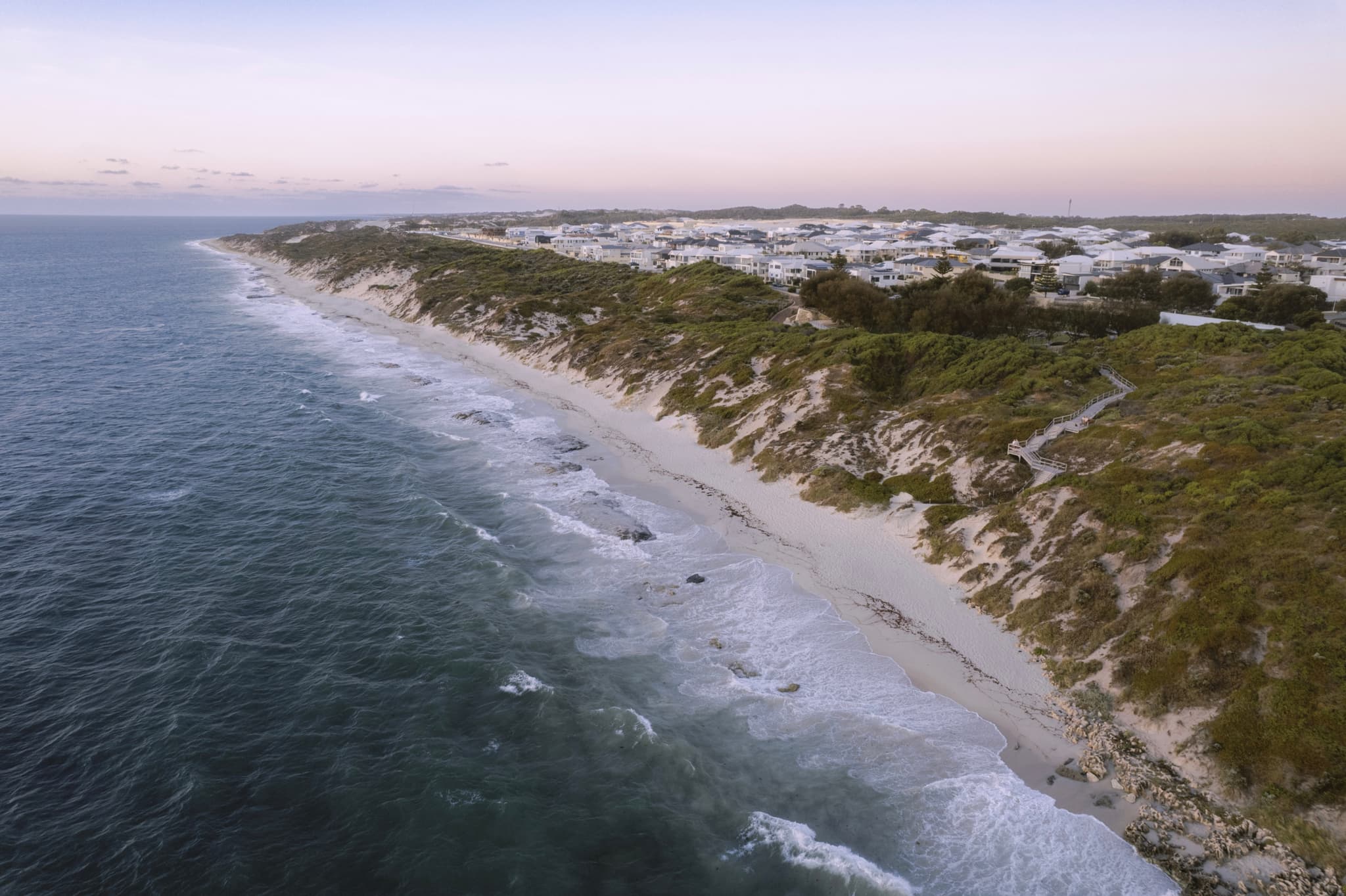 Aerial view of a coastal landscape with a sandy beach, waves crashing against the shore, and a line of white buildings atop a green cliff