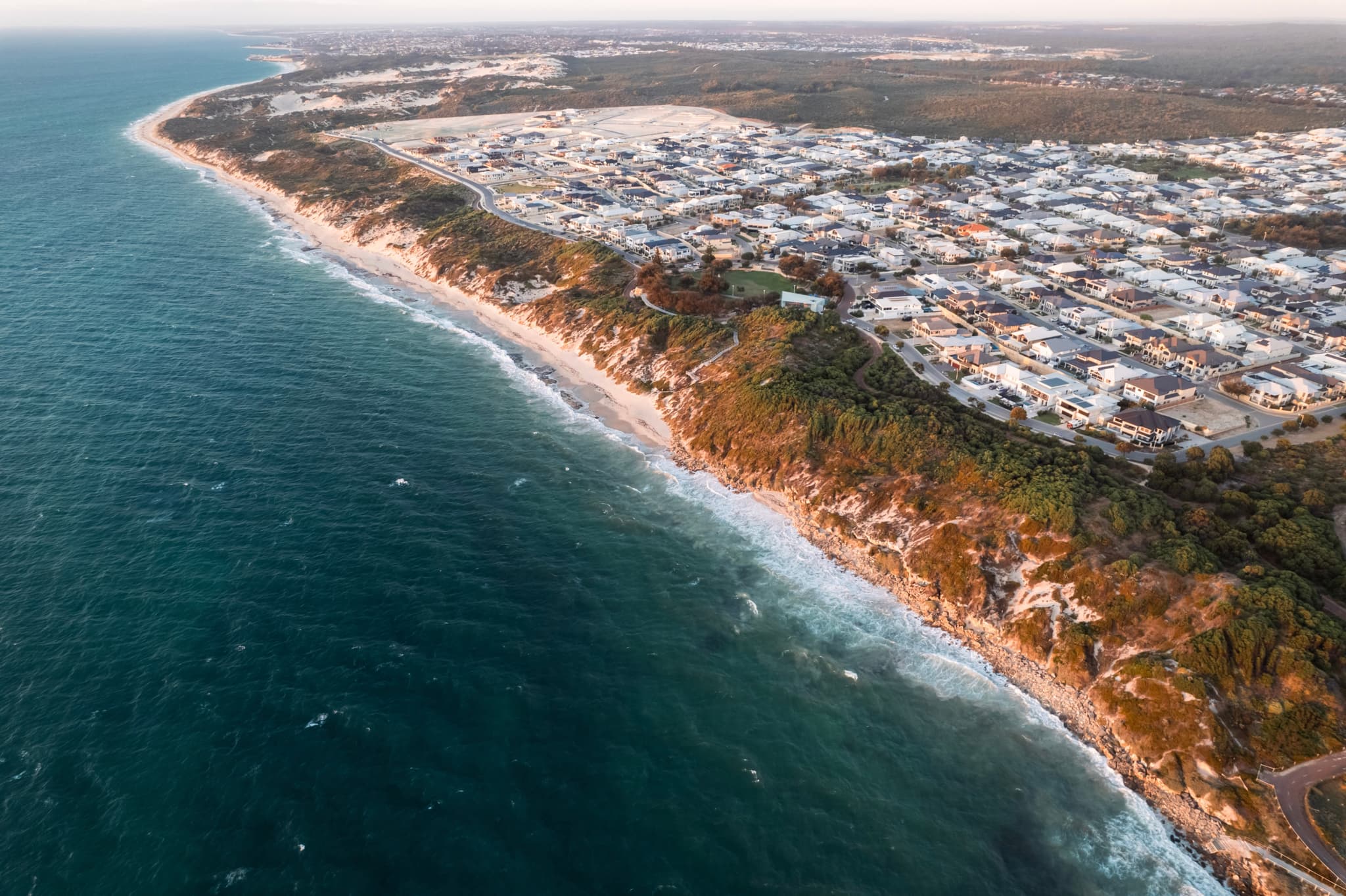 Aerial view of a coastal town with white buildings along a rugged shoreline, bordered by the ocean on one side and land on the other